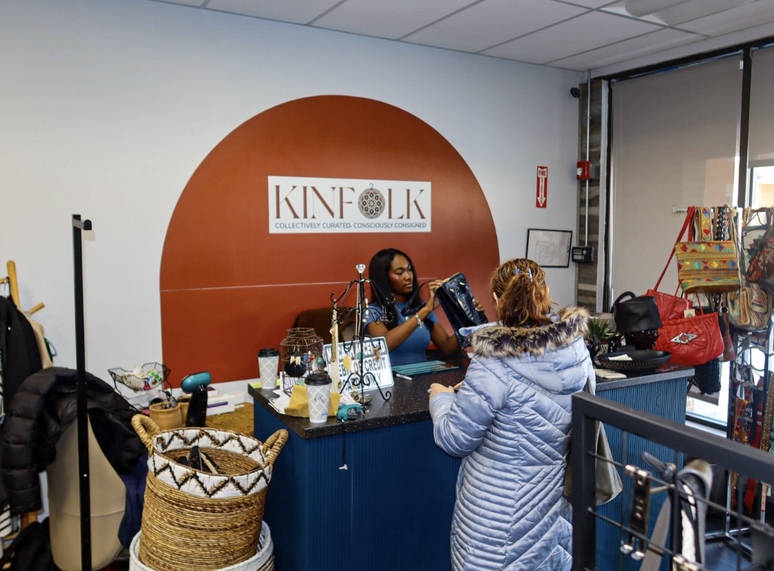 A Kinfolk Marketplace team member helping a shopper at the checkout counter, beneath the rust-arch Kinfolk logo wall