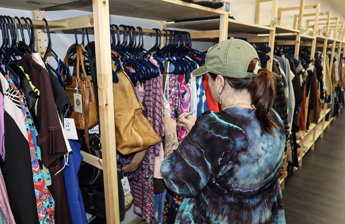 A shopper carefully browsing a wood-shelf rack of vintage clothing and leather handbags at Kinfolk Marketplace